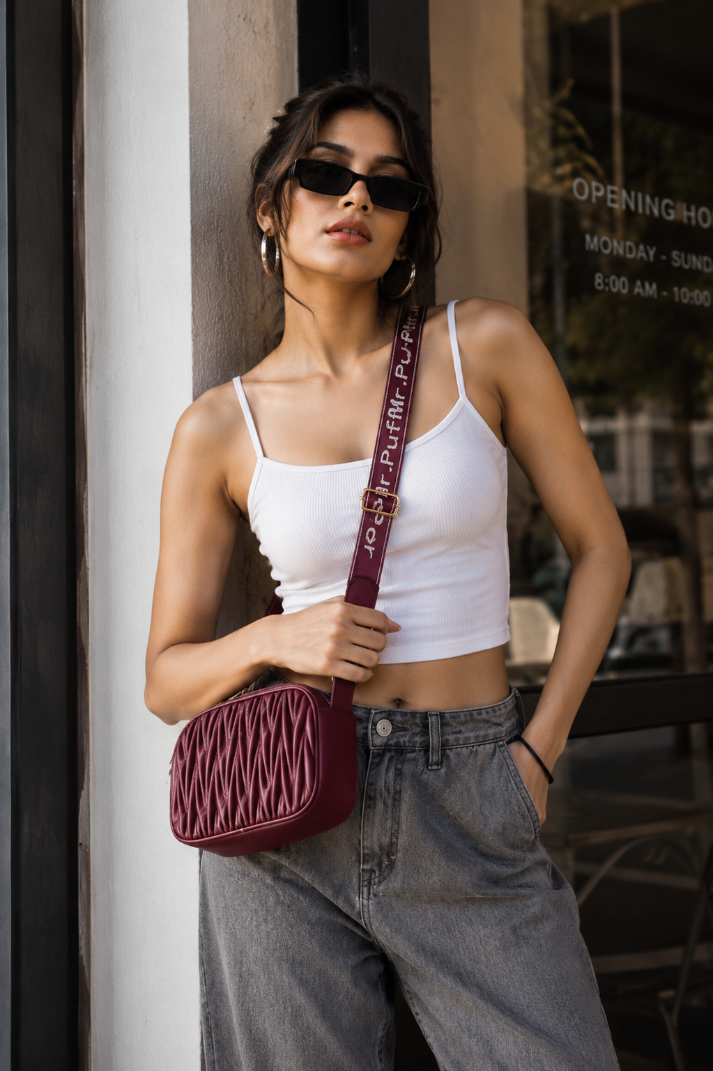 A woman wearing a white crop top and gray jeans models a burgundy quilted crossbody bag with a branded strap, standing in front of a storefront window.