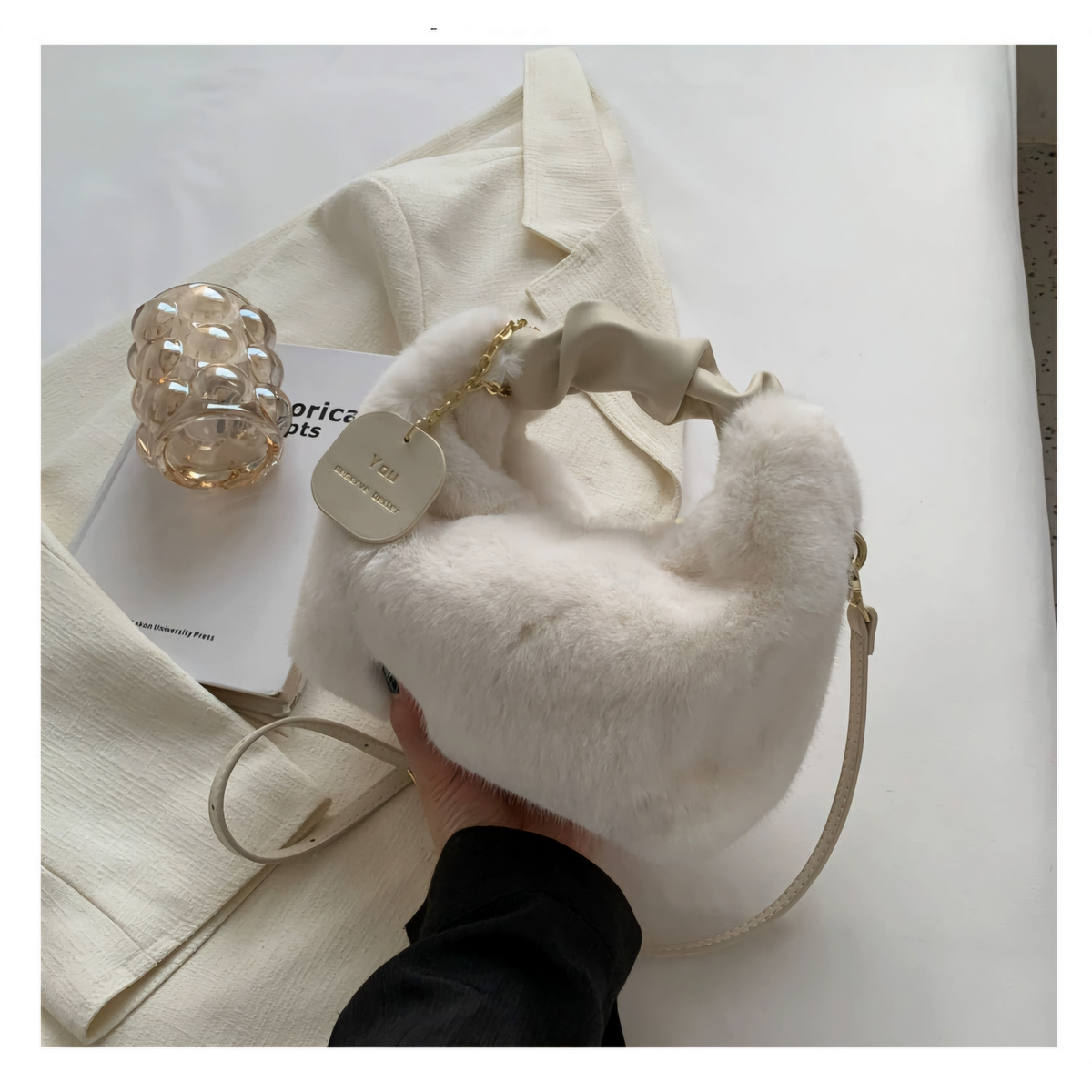 A fluffy off-white shoulder bag with a ribbon handle and gold chain detail, held by a hand over a cream-colored blazer. A small card tag on the bag reads 'MIA PARIS'.