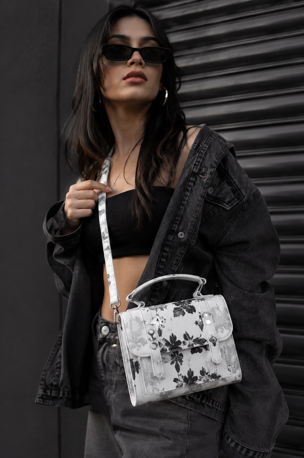 A woman wearing a black denim jacket, black crop top, and black sunglasses carries a white textured handbag with a black floral pattern and a silver handle, set against a dark corrugated metal background.