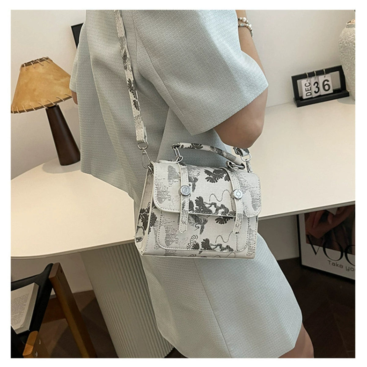 A person wearing a light gray dress carries a small structured shoulder bag with a white base and black floral print. The bag has a top handle, a shoulder strap, and silver-tone hardware, shown in an indoor room setting.