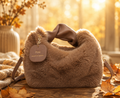 A soft brown faux-fur handbag with a wide brown handle and a hanging round tag reading 'You observe better,' styled on a wooden surface with autumn leaves and a blanket in a warm sunlit indoor setting.
