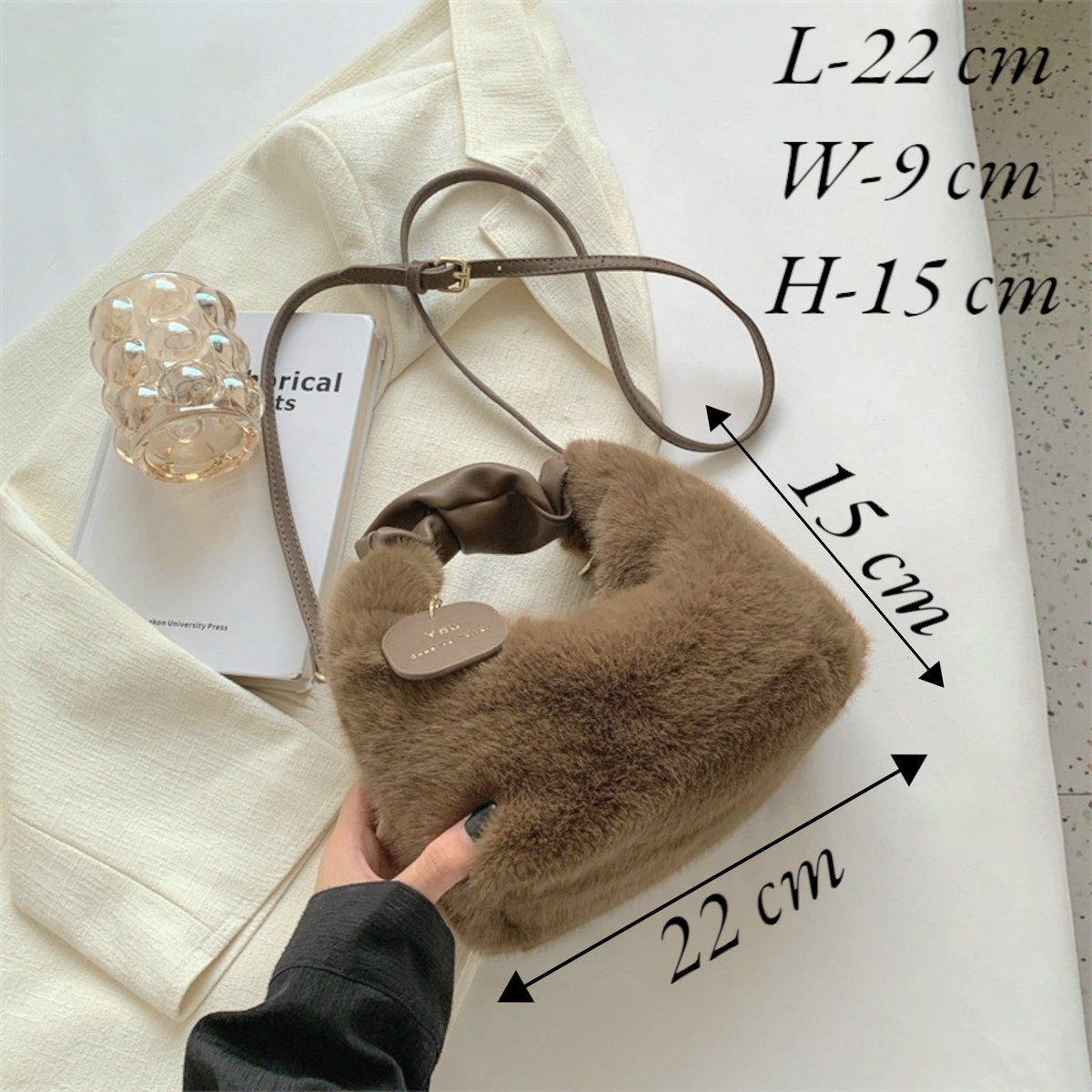 A brown fuzzy crescent shoulder bag with a brown strap, held by a hand over a cream fabric backdrop with overlaid size measurements and text showing dimensions.