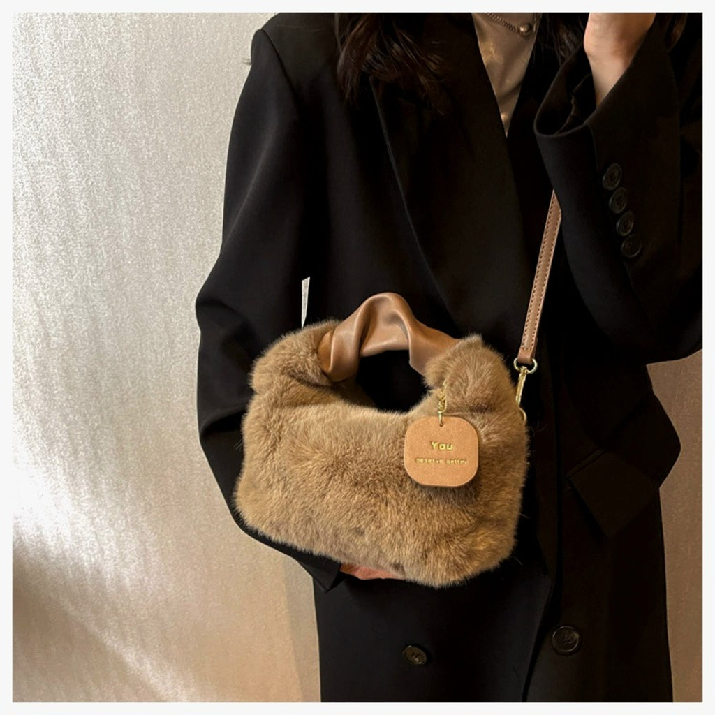 A person wearing a black blazer carries a small fuzzy shoulder bag in warm tan/beige. The bag has a smooth satin-like top handle, a thin matching strap, and a hanging tan tag with gold text that appears to read 'Velo' and 'Velo Travel Bag.
